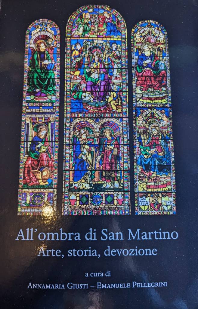 Book cover with a black background and a stained glass window. Text reads: All'ombra di San Martino: Arte, storia, devozione a cura di Annamaria Giusti - Emanuele Pellegrini
